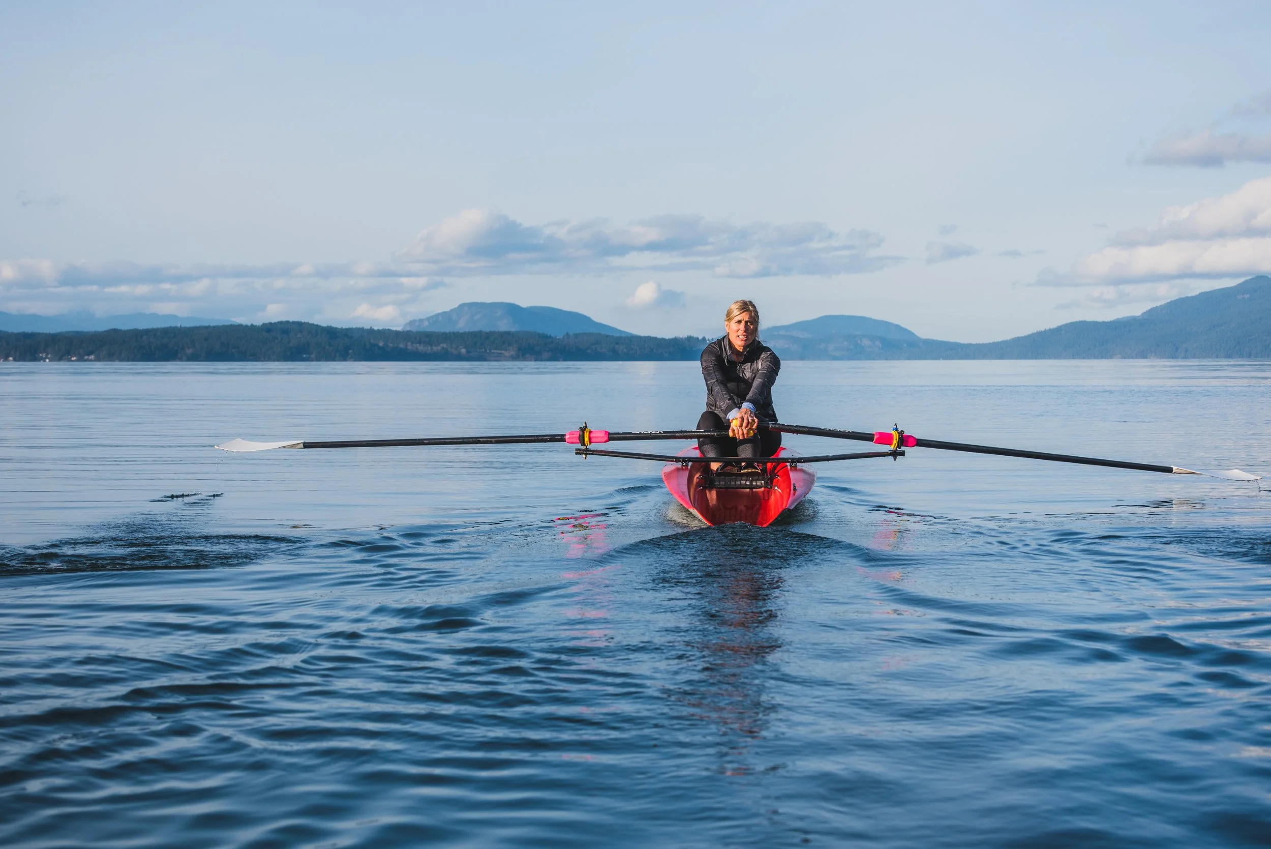 Silken Laumann resting at the oars of a single scull on flat water, mountains behind her